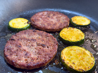 Close up of frying burger patties with zucchini slices in hot oil, homemade cooking of meat and vegetables on stove.