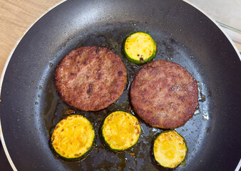 Fried zucchini slices and burger patties in pan with oil, close up view, homemade cooking process of meat and vegetables on stove.