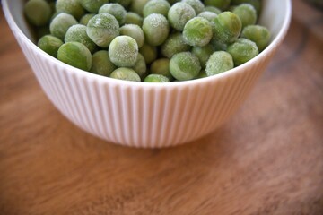 Fresh frozen green peas served in a white bowl on a rustic wooden table, close-up with copy space.