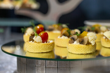 Delicate mini tartlets with mushrooms, cauliflower, cherry tomatoes, and cheese on a mirrored stand. Stylish appetizers for events, banquets, or fine dining.