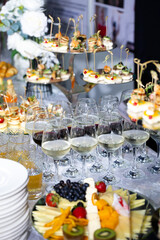 Close-up of a festive buffet table with glasses of red and white wine, assorted canapes on trays, and a fruit and cheese plate. Elegant catering setup for celebration, wedding or corporate event.