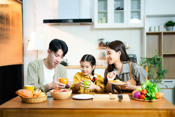 Smiling Asian family with daughter preparing fruits and vegetables together in a modern kitchen....