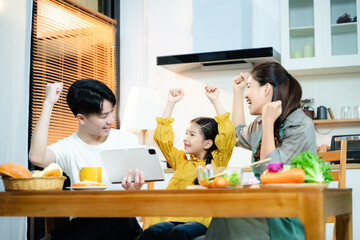 Happy Asian family cheers while using a tablet in the kitchen. Concept of digital learning, online success, family bonding, parenting, and joyful home moments.