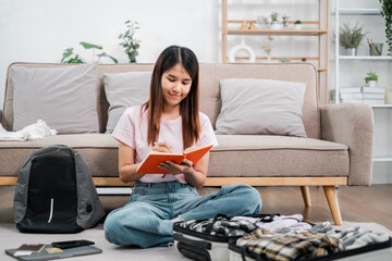 Obraz premium Smiling woman preparing for a trip, writing in a notebook, surrounded by travel essentials in a cozy living room.