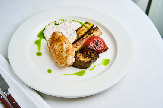 A platter features grilled vegetables including zucchini, tomato, and eggplant, served with a creamy dip and a warm bread roll. The dish is presented beautifully on a white background