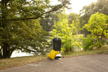 Overflowing trash bin with yellow bag and litter on the ground in a park near a lake.