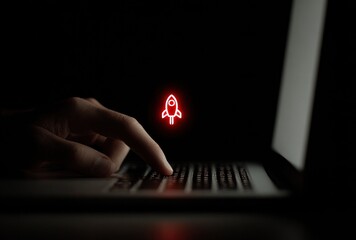 Hand types on a laptop in the dark, with a glowing red rocket icon floating above the keyboard. Focus is on the glowing launch symbol
