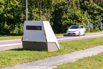 A speeding car is about to drive past a mobile road safety camera placed on the side of a busy road for traffic speed enforcement in a small town in France.