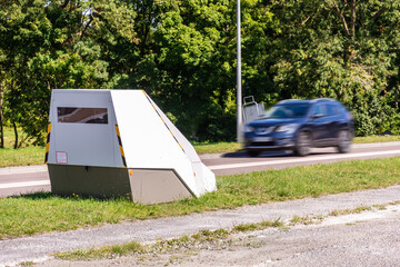 A speeding car is about to drive past a mobile road safety camera placed on the side of a busy road for traffic speed enforcement in a small town in France.