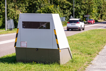 Cars drive past a mobile road safety camera placed on the side of a busy road for traffic speed enforcement in a small town in France.