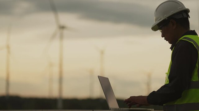 Engineer controlling smart grid system at wind farm using laptop. Renewable energy site management and clean power system monitoring technology in action.