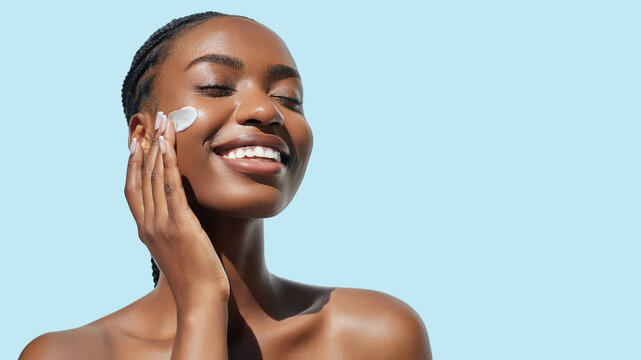 Overjoyed African American young woman with afro braids hairstyle is applying cosmetic product on her face Skin care beauty portrait. Cream smear. Hydration, nourishing and moisturising.