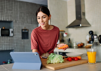 Happy young latina woman having fun preparing food and looking for recipes online using a tabletor laptop in kitchen