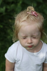 A blonde child in a white T-shirt stands in front of green leaves. The little girl squints from the sun. Portrait of a little blonde girl squinting.