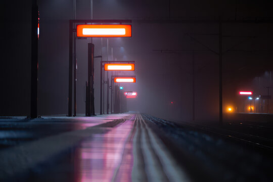 Illuminated train station platform at night with a train approaching in the distance and fog