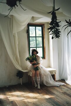 Teen girl in a white dress holding a bouquet, sitting by a window in a bright indoor setting.