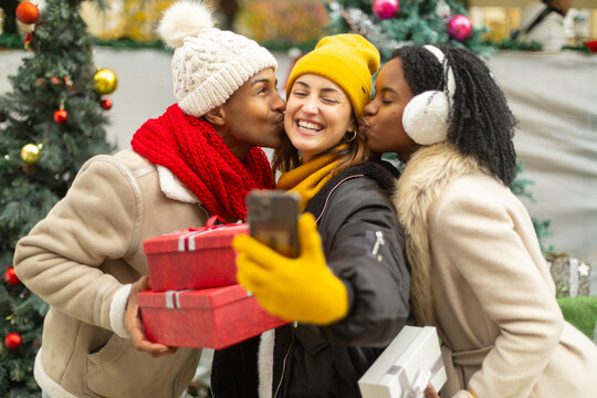Friends taking christmas selfie while kissing cheeks and holding gifts