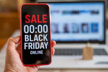Hand holding smartphone displaying black friday online sale countdown, with laptop featuring shopping website in background, highlighting online shopping and discounts