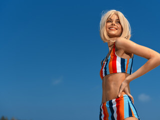 confident young woman wearing colorful striped swimsuit smiling happily, standing outdoors against clear blue sky, enjoying warm sunny day, summer vibes
