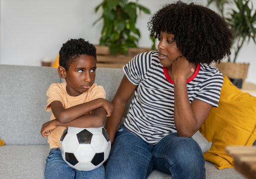 Sad boy holding soccer ball and sitting with mother on sofa at home
