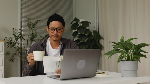 Asian man reading a book while drinking coffee at his home office desk. - Powered by Adobe