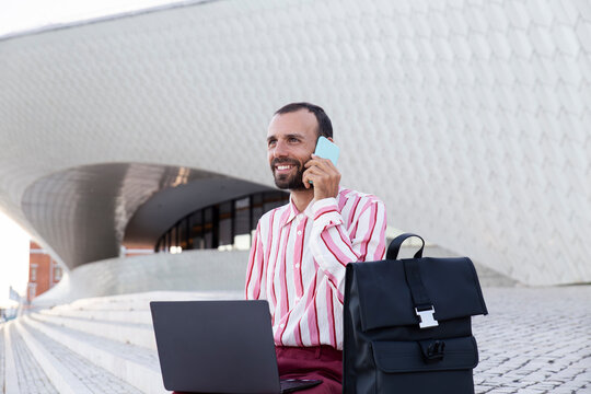 Smiling person having phone call with laptop and backpack outside Maat Lisbon
