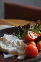 Close up of fresh burrata cheese served with ripe cherry tomatoes and salad leaves on a rustic wooden plate. Traditional Italian appetizer, gourmet dairy product, organic farm-to-table healthy food.
