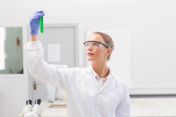 Scientist wearing lab coat, goggles and gloves holding green liquid test tube by microscope in lab