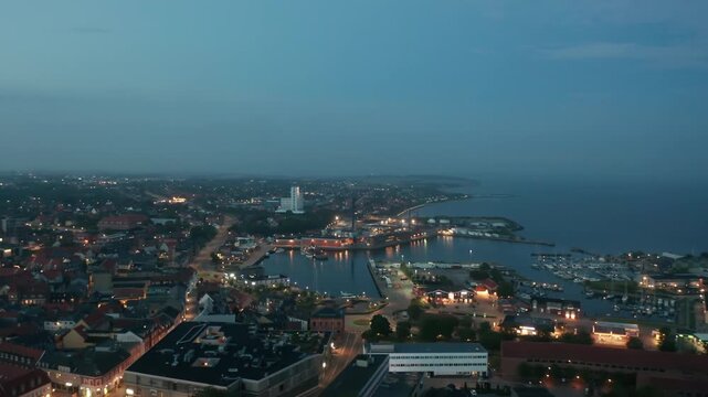 Aerial night view of Thisted, Denmark. Town lights, marina, Limfjord coastline, and surrounding neighborhoods create a tranquil Nordic cityscape under a twilight sky