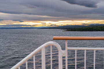 Orange and yellow hues fill the sky as the sun sets over Oslofjord in Vestfold County Norway. Dark clouds loom above the calm water, captured from a cruise ship railing.
