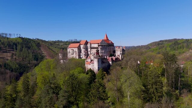 Slow aerial drone orbit around the towering Pernstein Castle, revealing its Gothic and Renaissance stone architecture amidst the green spring forest of South Moravia, Czechia.