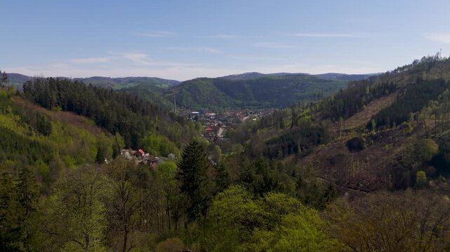 Aerial drone flyby past the iconic tower of Pernstein Castle, revealing the lush green valley and the historic town of Nedvedice in South Moravia, Czechia, under a clear blue spring sky.