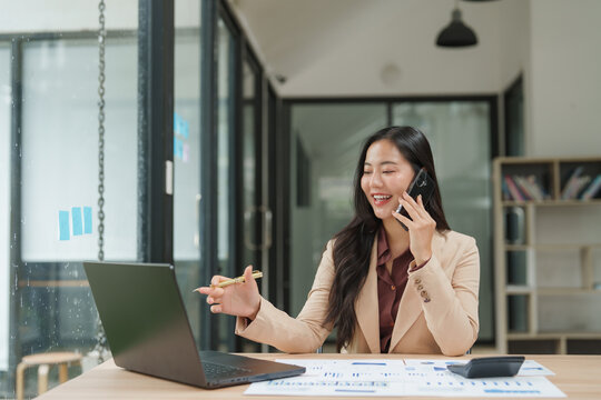 Asian businesswoman talking on phone and pointing at laptop in office - Powered by Adobe