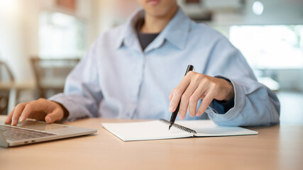Close up man holding pen writing in notebook and looking at laptop while sit at wooden table in cafe