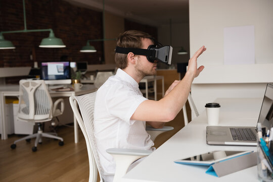 Male office worker wearing VR headset sitting at desk in open-plan office raising hand with laptop