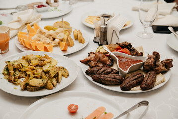 On a banquet table covered with a white tablecloth are various dishes of meat, vegetables, and fruits on white plates