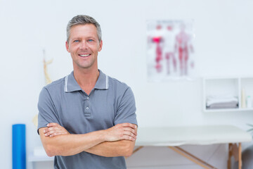 Mature male therapist standing arms crossed in therapy room next to treatment table