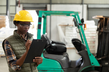 Woman wearing safety gear inspecting warehouse inventory using clipboard near forklift, copy space © wavebreak3