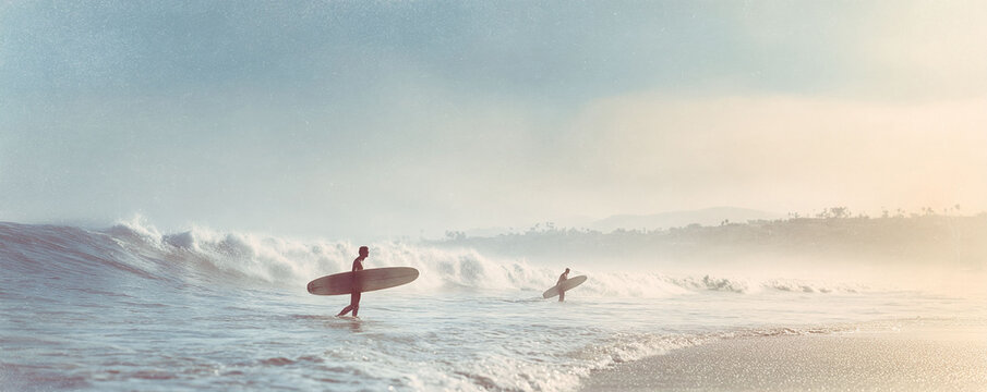 Silhouetted surfers enter the ocean with surfboards, facing large waves. Warm, vintage aesthetic. Evokes adventure, freedom,  the surfing lifestyle. Perfect for travel.