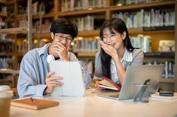 Fototapeta premium Glasses asian man student and woman friend looking and laughing at tablet at wooden table in library