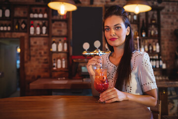 Woman in twenties holding orange-red cocktail with straw at bar counter with brick wall, copy space