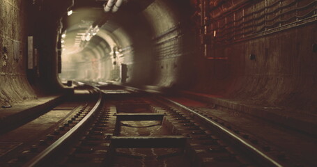 An underground railway tunnel features rusted tracks extending into darkness. Dim lights create an eerie atmosphere as shadows play along the walls. Water drips occasionally. © icetray