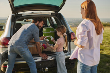 Family members gather around the open trunk of their car, unloading picnic supplies and smiling as they prepare for an enjoyable meal outdoors under the blue sky.
