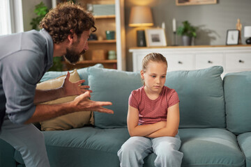 Obraz premium A father is talking to his daughter who looks upset while sitting on a couch at home. The atmosphere hints at a recent disagreement, with clear signs of tension in their expressions.