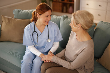 An elderly woman receives home care from a friendly, young care worker. They sit together on a comfortable sofa.