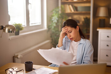 A woman sits at a wooden desk in a well-lit room, visibly stressed as she examines financial documents. She holds her head in frustration, surrounded by bills and a coffee cup.