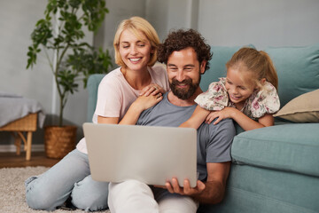 A family of three sits on a rug in a living room, engaging with a laptop. The adults smile and interact with a young girl who giggles and leans close.