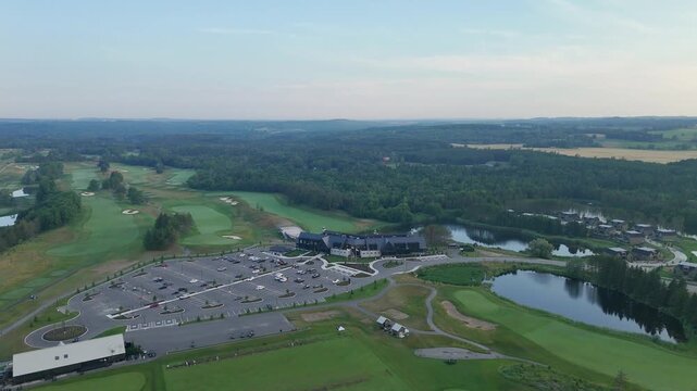 Drone orbiting aerial shot over TPC Toronto Osprey Valley golf course showing parking lot with vehicles, clubhouse, surrounding lakes, forest and golf fairways.