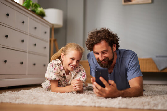 A father and his daughter are lying comfortably on a soft rug, laughing while they look at a smartphone together in a warm and inviting living room. - Powered by Adobe