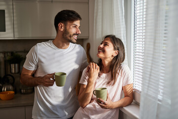 A couple stands in their cozy kitchen, holding mugs of coffee and smiling at each other, enjoying a peaceful moment together as sunlight streams through the window.